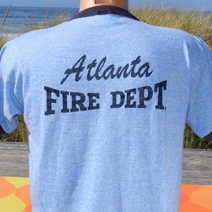 May include: A light blue ringer t-shirt with dark blue trim on the collar and sleeves. The back of the shirt features the dark blue text Atlanta FIRE DEPT printed in a combination of script and block lettering across the upper back area.