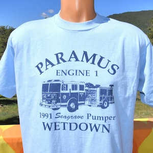 May include: Light blue t-shirt with a navy blue graphic of a fire truck and text. The text reads "PARAMUS ENGINE 1 1991 Seagrave Pumper WETDOWN". The shirt has short sleeves and a crew neck.