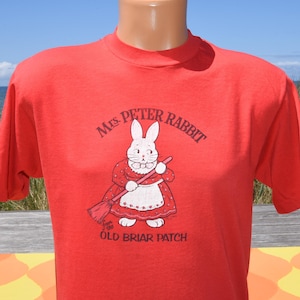 May include: A bright red short-sleeved t-shirt featuring a graphic of a white rabbit wearing a red dress and apron while holding a broom. The text reads Mrs. Peter Rabbit from the OLD BRIAR PATCH in black lettering above and below the rabbit.
