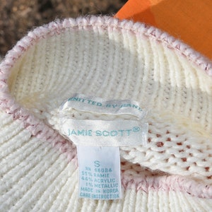 May include: White knitted sweater with a pink trim. The sweater has a label that reads "JAMIE SCOTT&reg;" and includes the size "S", RN number, fabric content, and care instructions.