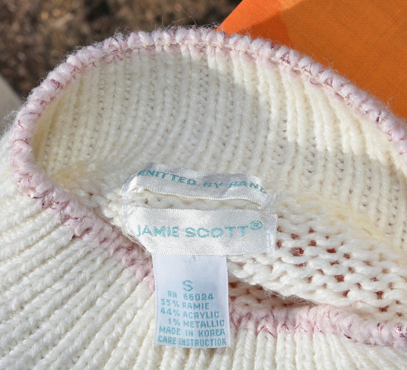 May include: White knitted sweater with a pink trim. The sweater has a label that reads "JAMIE SCOTT&reg;" and includes the size "S", RN number, fabric content, and care instructions.