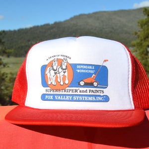 Op de afbeelding: Een rode en witte trucker-pet met een logo voor Superstriper en Paints. Het logo toont een team van Shire-paarden die een rode grasmaaier trekken met de tekst "Dependable "Workhorse"".
