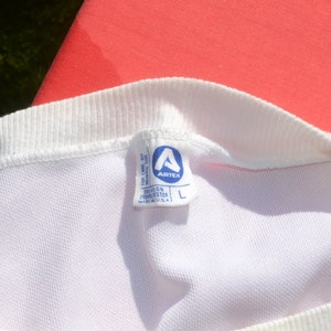 Może przedstawiać: Biała metka z niebieskim tekstem i niebieskim logo. Metka zawiera napis "Artex" i "Teflon 100% Polyester Made in U.S.A." z etykietą rozmiaru "L".