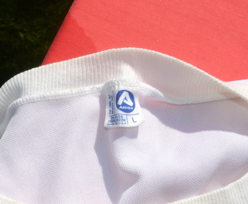 Może przedstawiać: Biała metka z niebieskim tekstem i niebieskim logo. Metka zawiera napis "Artex" i "Teflon 100% Polyester Made in U.S.A." z etykietą rozmiaru "L".