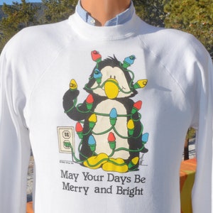 May include: White sweatshirt featuring a cartoon penguin wrapped in colorful Christmas lights. The penguin has a yellow beak and feet. The text "May Your Days Be Merry and Bright" is printed below the graphic. The sweatshirt has long sleeves.