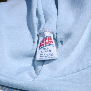 以下が含まれることがあります： 「SOFFE SWEATS」と「ADULT XL 46-48」と書かれたタグが付いた、水色のパーカーのクローズアップ。タグには、パーカーがアメリカ合衆国ノースカロライナ州フェイエットビルで製造されたことも記載されています。パーカーは柔らかい素材で作られています。
