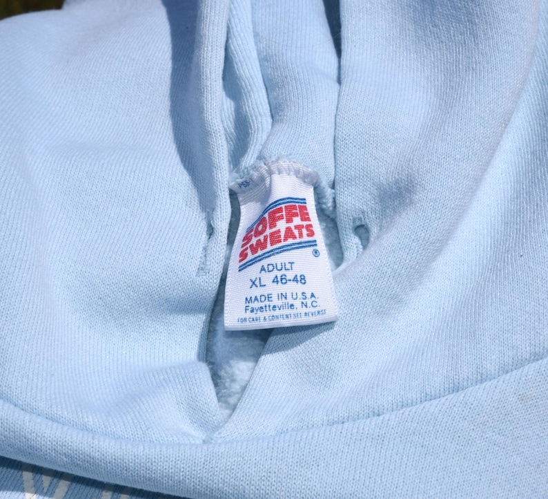 以下が含まれることがあります： 「SOFFE SWEATS」と「ADULT XL 46-48」と書かれたタグが付いた、水色のパーカーのクローズアップ。タグには、パーカーがアメリカ合衆国ノースカロライナ州フェイエットビルで製造されたことも記載されています。パーカーは柔らかい素材で作られています。