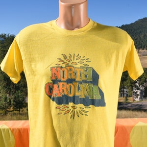 Pu&ograve; includere: Maglietta gialla a maniche corte con una stampa vintage di "NORTH CAROLINA" in lettere colorate con un design a raggiera.