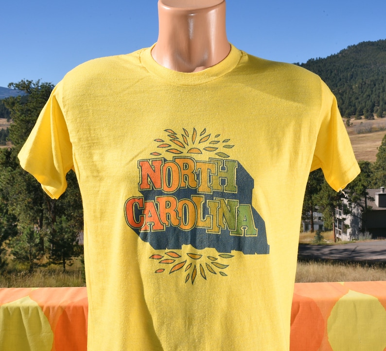 Pu&ograve; includere: Maglietta gialla a maniche corte con una stampa vintage di "NORTH CAROLINA" in lettere colorate con un design a raggiera.