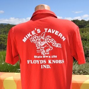 May include: Red short-sleeved polo shirt with a white graphic on the back. The graphic features the text "MIKE'S TAVERN BAR HOPPER" and a cartoon rabbit holding a drink. Below the graphic is "State Hwy. 150 FLOYDS KNOBS IND."
