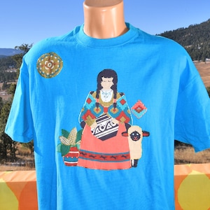 May include: A turquoise blue t-shirt with a colorful embroidered design of a woman in traditional clothing holding a pot and a lamb. The design also includes a small embroidered flower pot with corn stalks.