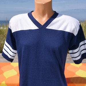 May include: A short-sleeve V-neck shirt in navy blue with a white panel across the upper chest and shoulders. The sleeves feature three horizontal white stripes near the cuffs, creating a classic athletic-inspired design on this cotton-blend top.