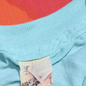 May include: Close-up of a light blue knit shirt with a tag. The shirt has a ribbed neckline and appears to be made of a soft, breathable fabric. The tag is off-white with faded text and graphics.