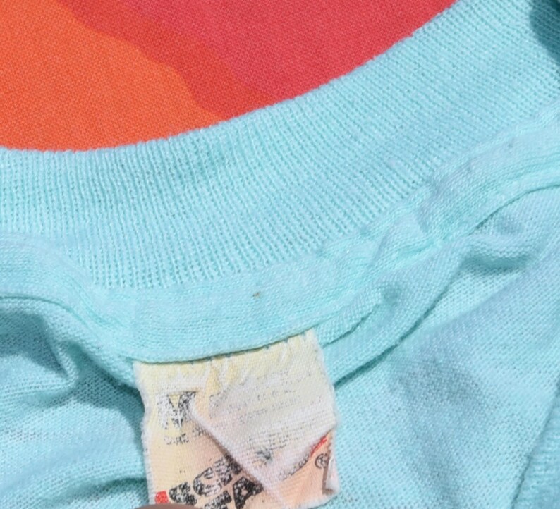 May include: Close-up of a light blue knit shirt with a tag. The shirt has a ribbed neckline and appears to be made of a soft, breathable fabric. The tag is off-white with faded text and graphics.