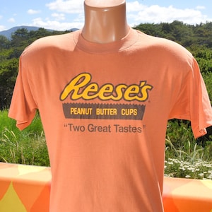 May include: Orange t-shirt featuring the Reese's Peanut Butter Cups logo in yellow and brown. The shirt also includes the text "Two Great Tastes" below the logo. The t-shirt is displayed on a mannequin.