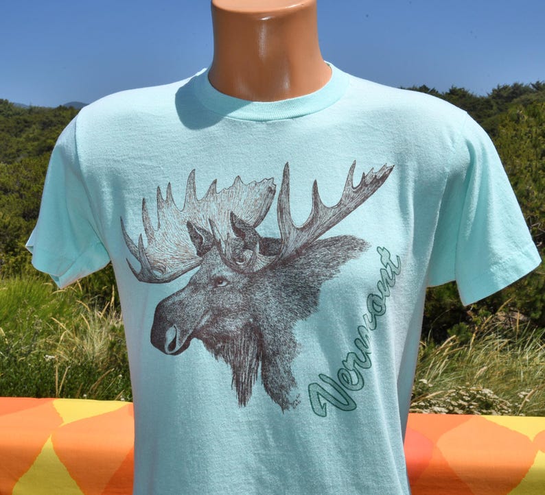 May include: A light blue t-shirt featuring a detailed illustration of a moose head with large antlers. The word "Vermont" is written in a script font on the right side of the moose. The shirt is displayed on a mannequin.