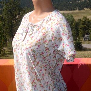 May include: White sheer blouse with a floral print featuring pink, blue and orange flowers. The blouse has a tie at the neckline.