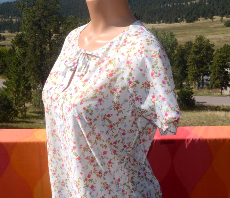 May include: White sheer blouse with a floral print featuring pink, blue and orange flowers. The blouse has a tie at the neckline.