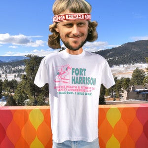 May include: White t-shirt with a pink and green graphic of a person running. The graphic includes the text "FORT HARRISON", "EMPLOYEE HEALTH & FITNESS DAY", "SAFETY AWARENESS DAY", and "2 MILE RUN - 1 MILE WALK".
