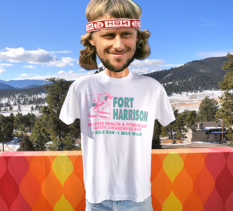 May include: White t-shirt with a pink and green graphic of a person running. The graphic includes the text "FORT HARRISON", "EMPLOYEE HEALTH & FITNESS DAY", "SAFETY AWARENESS DAY", and "2 MILE RUN - 1 MILE WALK".