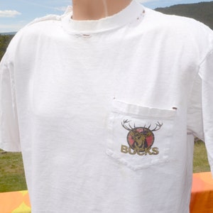 Op de afbeelding: Wit T-shirt met een verweerde look en een zakje aan de linkerkant. Het zakje heeft een afbeelding van een hertenkop met het woord "BUCKS" eronder.