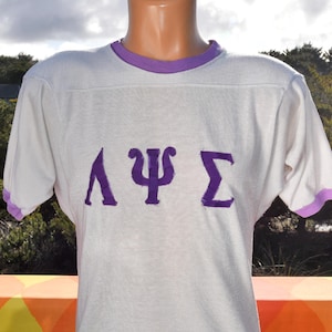 May include: A vintage white t-shirt with purple trim on the collar and sleeves. The shirt features three purple Greek letters across the chest. The shirt is made of a soft, possibly cotton, material.