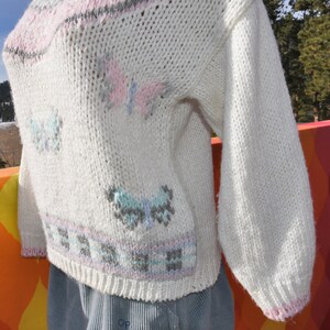 May include: White knitted sweater with a pink, blue, and gray butterfly design. The sweater has a ribbed neckline and cuffs.