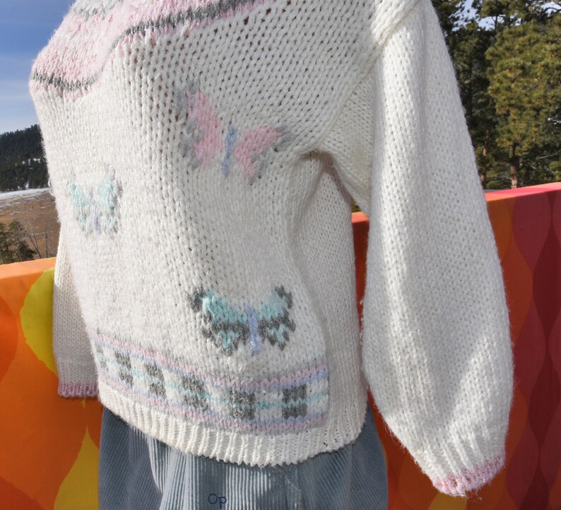 May include: White knitted sweater with a pink, blue, and gray butterfly design. The sweater has a ribbed neckline and cuffs.