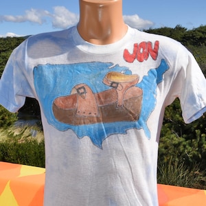 May include: White t-shirt featuring a hand-drawn design of a brown saddle on a blue map of the United States. The name "JON" is written in red above the map. The shirt has short sleeves.