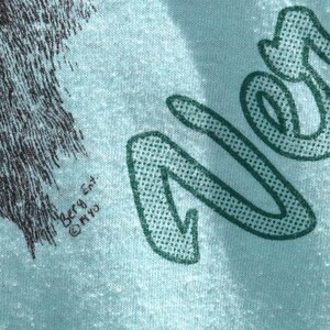 May include: Close-up of a light blue t-shirt with a graphic design. The design includes the word "Never" in a green, dotted font. The shirt also features a black and gray illustration of a wolf. The text "Berg Ent. &copy; MYO" is printed on the shirt.