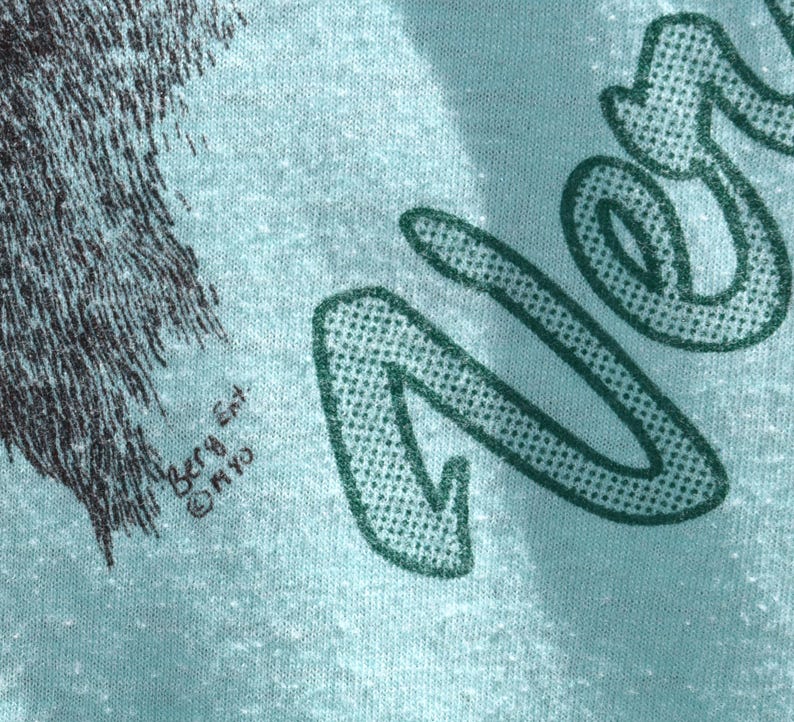 May include: Close-up of a light blue t-shirt with a graphic design. The design includes the word "Never" in a green, dotted font. The shirt also features a black and gray illustration of a wolf. The text "Berg Ent. &copy; MYO" is printed on the shirt.