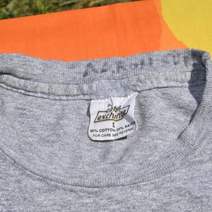 May include: Close-up of a heather gray t-shirt with a white tag. The tag reads "the cotton exchange" and includes the fabric composition: 90% cotton, 10% rayon. The shirt is size small.