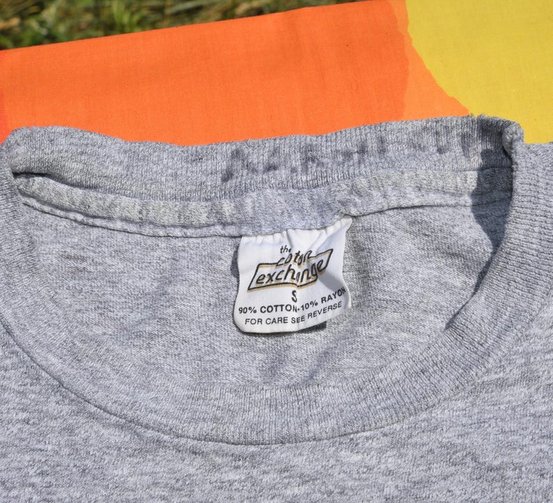 May include: Close-up of a heather gray t-shirt with a white tag. The tag reads "the cotton exchange" and includes the fabric composition: 90% cotton, 10% rayon. The shirt is size small.