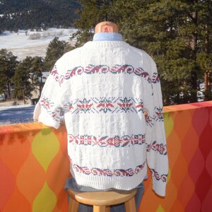 May include: White knitted sweater with a colorful geometric pattern. The sweater has a crew neck and long sleeves.