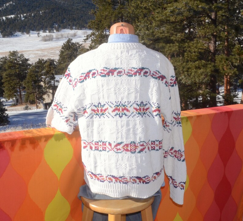 May include: White knitted sweater with a colorful geometric pattern. The sweater has a crew neck and long sleeves.