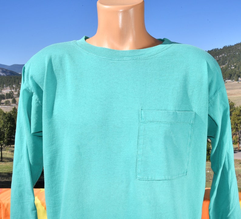 May include: A turquoise long-sleeved t-shirt with a single pocket on the left side.