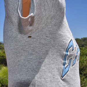 May include: A gray sleeveless shirt with a blue and white graphic on the side. The shirt is made of a soft material and has a relaxed fit. The graphic appears to be a stylized letter or symbol.