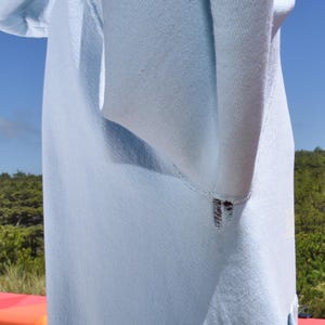 以下が含まれることがあります： 水色のフード付きスウェットシャツのクローズアップ。生地の質感と縫い目を示しています。画像は、衣類の構造を強調しています。背景には青い空と緑の葉があります。