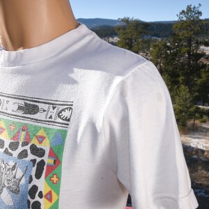 May include: White t-shirt featuring a colourful graphic print of a rhinoceros and geometric patterns. The design incorporates a blue, green, yellow, red, and white colour scheme.
