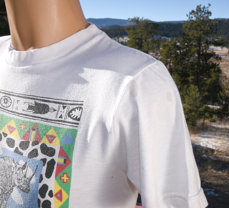 May include: White t-shirt featuring a colourful graphic print of a rhinoceros and geometric patterns. The design incorporates a blue, green, yellow, red, and white colour scheme.