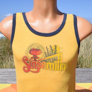 May include: Mustard yellow tank top with navy blue trim. The front features a graphic with the word "Sinsemilla" in red and yellow, along with a design of a stylized mushroom, trees, and a sun. The tank top is displayed on a mannequin.