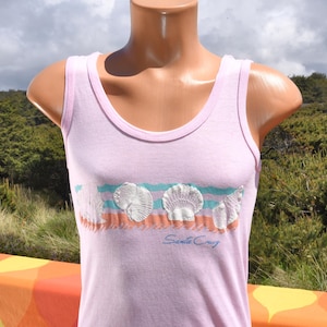 May include: A light purple tank top with a scoop neck and a seashell graphic. The graphic features white seashells, blue and orange stripes, and the text "Santa Cruz" in a script font. The tank top is displayed on a mannequin.