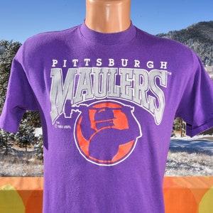 May include: Purple t-shirt with a white graphic of a football player wearing a helmet and holding a football. The graphic says "Pittsburgh Maulers" and "TM 1983 USFL".