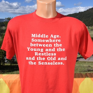 May include: A red t-shirt with white text that reads "Middle Age. Somewhere between the Young and the Restless and the Old and the Senseless." The shirt is displayed on a mannequin.