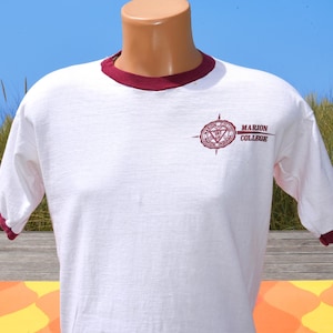 May include: A white short-sleeve ringer t-shirt featuring a maroon crew neckline and matching maroon sleeve cuffs. The chest displays a maroon graphic logo of a compass-style emblem with the text Marion College, Marion, Indiana printed beside it.