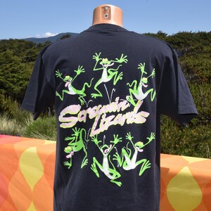 May include: Dark navy blue t-shirt featuring a design of green cartoon frogs with white accents. The text "Sorguin Lizards" is printed in a pink and yellow font. The shirt is displayed on a mannequin.