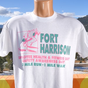 May include: White t-shirt with a pink and green graphic of two people running. The text "FORT HARRISON" is in green and pink. The text "EMPLOYEE HEALTH & FITNESS DAY SAFETY AWARENESS DAY 2 MILE RUN - 1 MILE WALK" is in black.