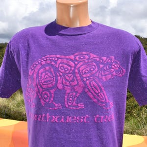 Puede incluir: Camiseta morada con un dise&ntilde;o de oso estilizado en rosa. El gr&aacute;fico del oso presenta intrincados patrones de inspiraci&oacute;n tribal. El texto "Northwest Tre" est&aacute; impreso debajo. La camiseta tiene mangas cortas y cuello redondo. La tela es suave.