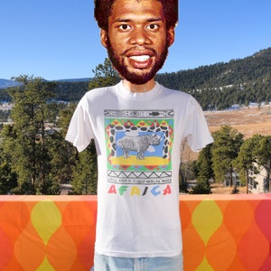 May include: White t-shirt featuring a colourful graphic of a rhinoceros within a patterned frame. The text "AFRICA" is printed below the graphic. The text "NATIONAL PARKS - HIGHLANDS WILDLIFE AREAS" is printed above the graphic.