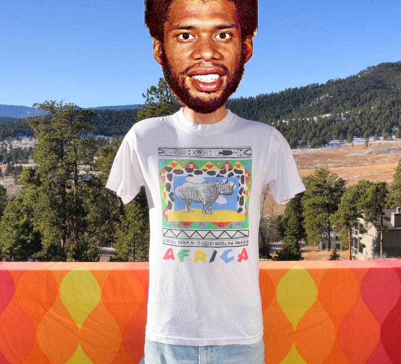 May include: White t-shirt featuring a colourful graphic of a rhinoceros within a patterned frame. The text "AFRICA" is printed below the graphic. The text "NATIONAL PARKS - HIGHLANDS WILDLIFE AREAS" is printed above the graphic.